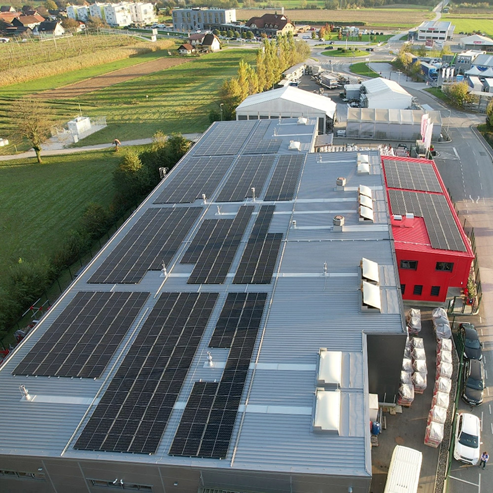 Zračni posnetek industrijskega objekta z večjimi polji sončnih panelov na kovinski ravni strehi; v ozadju poslovna cona in parkirišče (GOMARK d.o.o.).